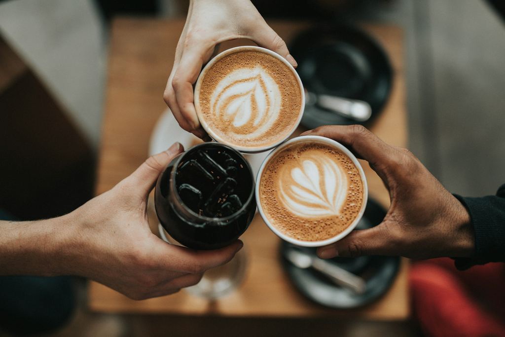 Three hands seen from above, raising two lattes with leaf-patterned foam art and a glass of cold brew over a wooden table