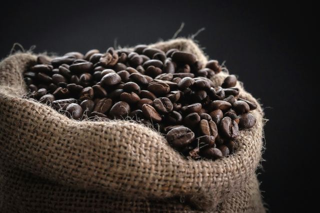 Dark-roasted coffee beans heaped in an open burlap sack against a black background